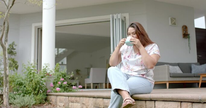 Happy plus size biracial woman sitting on garden terrace drinking coffee, copy space, slow motion