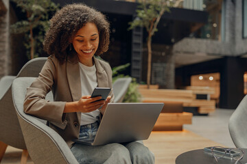 Positive female freelancer holding mobile phone while sitting in coworking and work on laptop