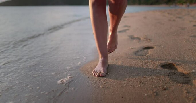 Barefoot legs walking sea shore waves, summer day. Woman feet leave footprints at sand beach, sunset outdoors. Female enjoying resting on summer vacation. Travel, tourism, journey concept. Slow motion