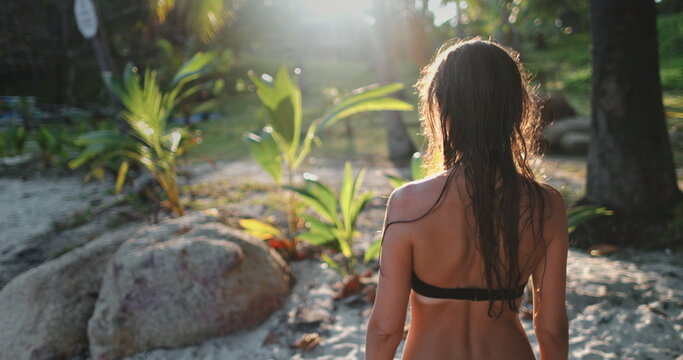 Woman in bikini walking sunset tropical beach garden, soft light. Slim girl enjoy and relax makes her way in green palm leaves. Outdoor lifestyle travel summer holiday vacation. Back view