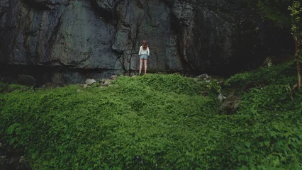 Wandaufkleber Aerial view tourist woman standing in green tropical jungle forest against high rock waterfall. Outdoor lifestyle travel on summer holiday vacation. Dramatic dark toning. Drone shot © Anastasia Pro