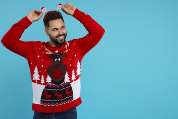 Happy young man in Christmas sweater and Santa headband on light blue background. Space for text