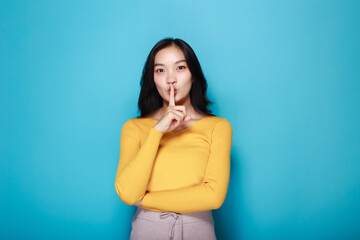 An Asian woman poses to signal silence while standing in front of a light blue background.