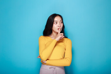 An Asian woman poses to signal silence while standing in front of a light blue background.