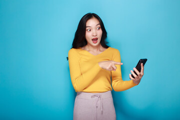 Naklejka premium Young happy woman showing point fingers isolated while holding a cell phone in the other hand, Standing and pointing happily, light blue back ground.