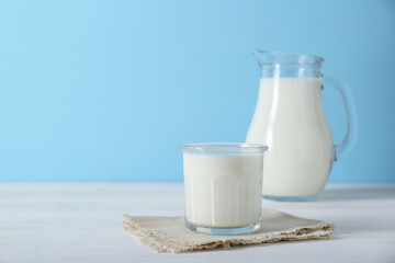 Glass of fresh milk and jug on white wooden table against light blue background, space for text