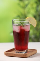 Refreshing hibiscus tea with ice cubes, mint and lemon in glass on white table against blurred green background