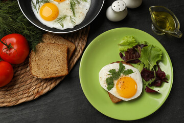 Frying pan and plate with tasty fried eggs, salad and products on black table, flat lay