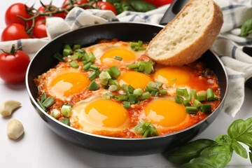 Delicious Shakshuka in frying pan and bread piece on white tiled table, closeup