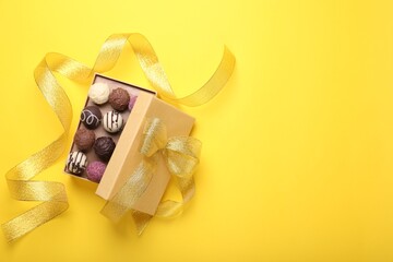 Open box with delicious chocolate candies and ribbon on yellow background, top view. Space for text