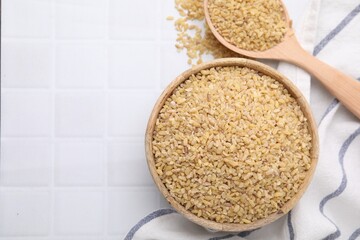 Bowl and spoon with raw bulgur on white tiled table, flat lay. Space for text