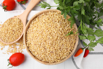 Raw bulgur in bowl, spoon, vegetables and parsley on table, flat lay