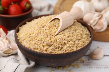 Bowl and scoop with raw bulgur on white wooden table, closeup
