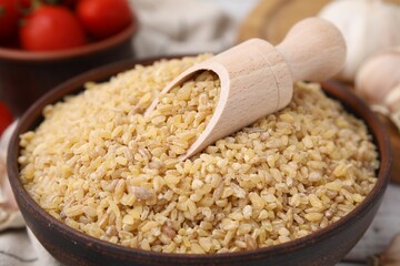 Bowl and scoop with raw bulgur on table, closeup