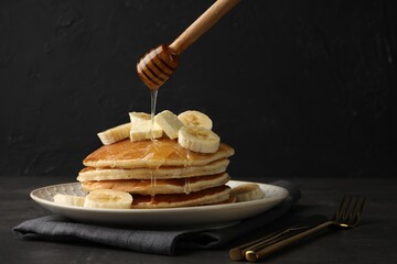 Pouring honey from dipper onto delicious pancakes with bananas and butter at dark table, space for...