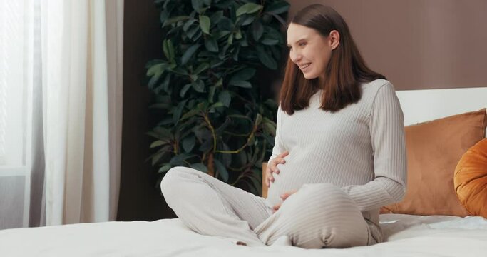 Expecting Joy concept happy smiley beautiful pregnant woman with a short hairstyle sits on her bed, lovingly touching her belly and smiling with anticipation for her little one's arrival.