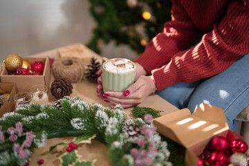 Naklejka na ściany i meble Young asian woman in sweater happiness and drinking hot chocolate with marshmallow after making christmas wreath to preparing ornaments for celebrate christmas festive holiday and winter seasons