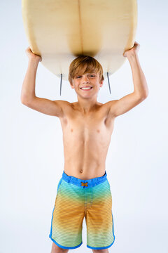 Smiling cute surfer boy in colorful board shorts carrying his surfboard over his head