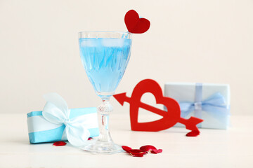 Glass of tasty cocktail with gift and rose petals on wooden table against white background. Valentine's Day celebration