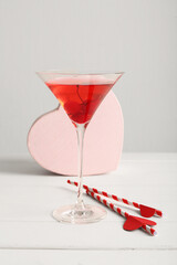 Glass of tasty cocktail with straws and decorative heart on wooden table against white background. Valentine's Day celebration