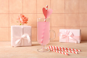 Glass of tasty cocktail with straws and gifts on table against beige tile background. Valentine's Day celebration