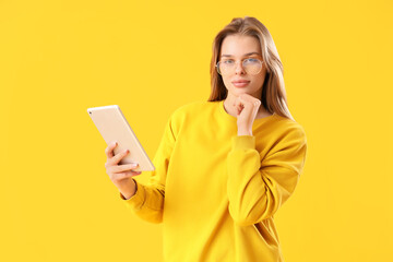 Thoughtful young woman in eyeglasses using tablet computer on yellow background