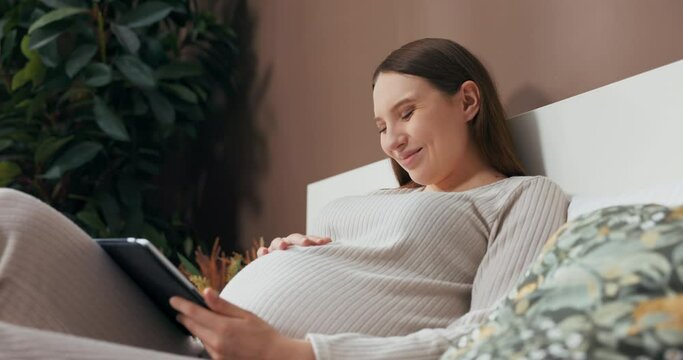 Dreamy alluring pregnant woman dressed in a stylish and comfortable outfit, sitting on a bed, holding a tablet, and admiring photos with interest.