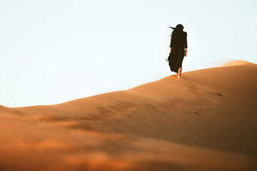 Low angle beautiful woman in long black dress barefoot walk up follow walk on golden Sahara desert...