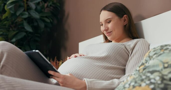 Delighted pregnant woman in a trendy and comfortable outfit sits on a bed, holding a tablet, and browsing through photos with a smile on her face.