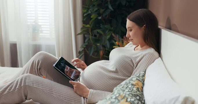 Attractive pregnant woman in a stylish and comfortable outfit sits on a bed holding a tablet, scrolling through photos with a curious expression.