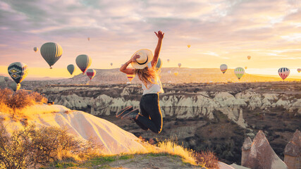 Young female tourist jumping in the air with hot air balloons background at sunrise in Cappadocia, Turkey - obrazy, fototapety, plakaty