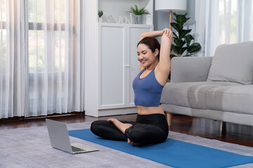Fototapeta na wymiar Asian woman in sportswear doing yoga exercise on fitness mat as her home workout training routine. Healthy body care lifestyle woman watching online yoga video on laptop. Vigorous