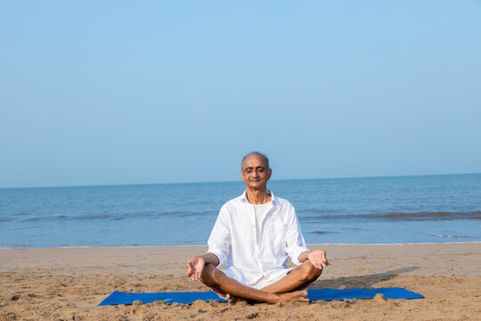 Senior indian Man sitting on fitness mat in lotus position meditating at beach. Healthy Life Concept.