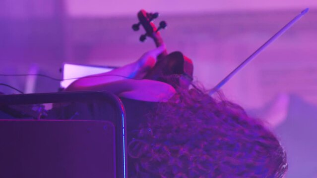 A violinist girl with curly hair plays the violin. View from behind. Sympho-rock band performing. Close-up of a woman's head and a violin. A blurred guitarist in the background