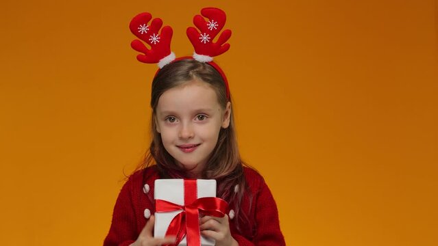 Happy child with the long-awaited gift. A girl in a red Christmas sweater and deer horns on her head presses a gift box to her cheek. The girl is happy that her dream has come true and thanks Santa 