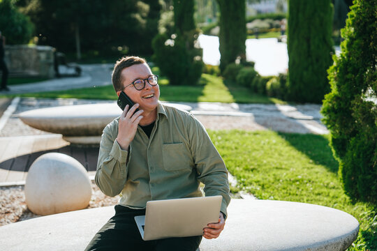 Cheerful young man in glasses and casual clothes sitting at public park with laptop talks by phone toothy smiles enjoying conversation with friends. Relocation, freelancer works outdoors.