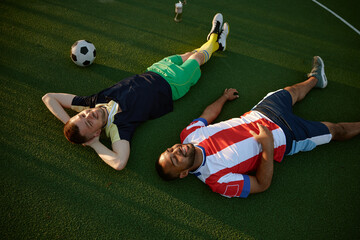 Smiling restful two football players lying on green field grass overhead view