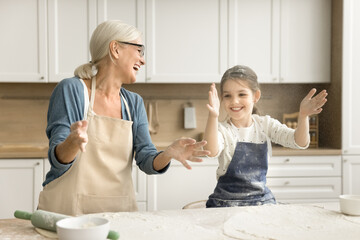 Fototapeta premium Excited grandmother and happy little granddaughter girl in aprons throwing flour over table with ingredients laughing, having fun, clapping floury hands for making cloud, playing while cooking