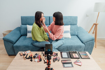 Focused ethnic women sitting on sofa with makeup kit and recording video - obrazy, fototapety, plakaty