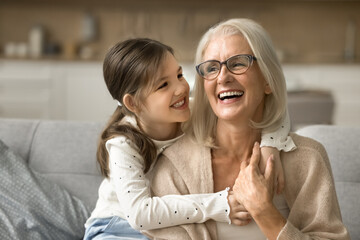 Happy blonde grandma and cheerful granddaughter child playing active games on home sofa. Grandmother and grandkid enjoying funny leisure on family meeting, having fun, hugging on couch - obrazy, fototapety, plakaty