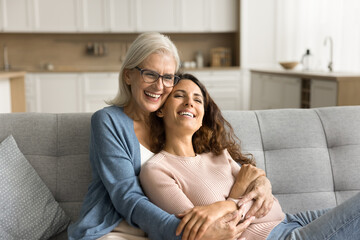 Joyful mature mother and adult child woman sitting together on sofa, hugging, laughing, talking,...