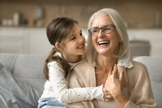 Happy blonde grandma and cheerful granddaughter child playing active games on home sofa. Grandmother and grandkid enjoying funny leisure on family meeting, having fun, hugging on couch