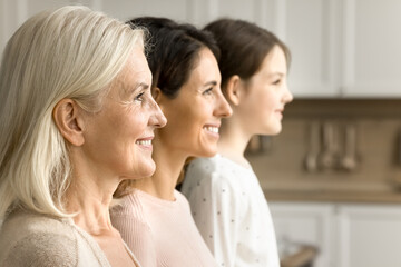 Cheerful blonde elderly grandmother looking forward away side shot with brunette daughter woman and grandkid girl standing in line in blurred background. Three female generations family portrait - obrazy, fototapety, plakaty