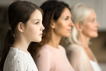 Side shot of serious little tween child girl standing in row with mom and grandmother in blurred background, looking forward away, posing for family portrait of three female generations
