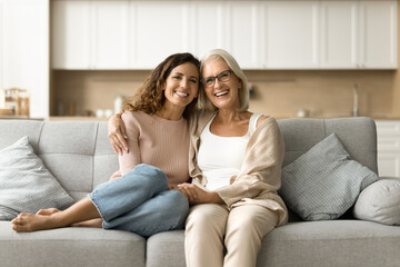 Cheerful senior mother and daughter woman sitting on home couch close together, looking at camera, smiling for portrait. Happy mature mom hugging adult kid, laughing, enjoying family leisure