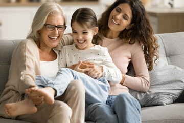Happy joyful grandma and mom playing with cheerful kid girl on sofa, tickling laughing child,...