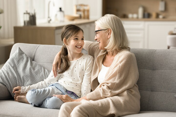 Cheerful loving grandmother and cute grandkid girl talking and laughing on sofa, enjoying funny conversation, trust, family leisure. Grandma speaking to child girl at home