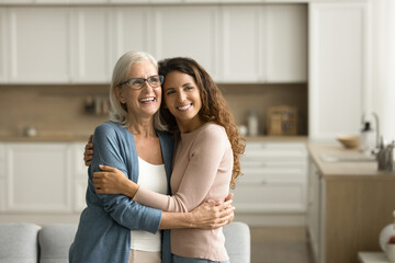 Cheerful dreamy elder mom and positive adult daughter woman hugging at home with love, support, looking away, smiling, thinking on family relationships, bonding, discussing mothers retirement plans
