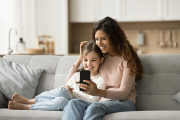 Cheerful pretty black haired mom and positive daughter child talking on video call on smartphone, resting on home couch, using mobile phone for online family communication
