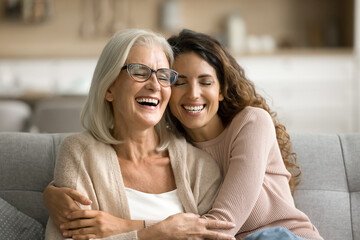 Happy excited senior mom and adult daughter woman having fun at home, hugging with heads touch, sitting on sofa, laughing with closed eyes, enjoying funny talk, leisure, family affection - obrazy, fototapety, plakaty